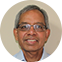A smiling older man with short gray hair and glasses, wearing a light blue collared shirt, pictured against a plain beige background.