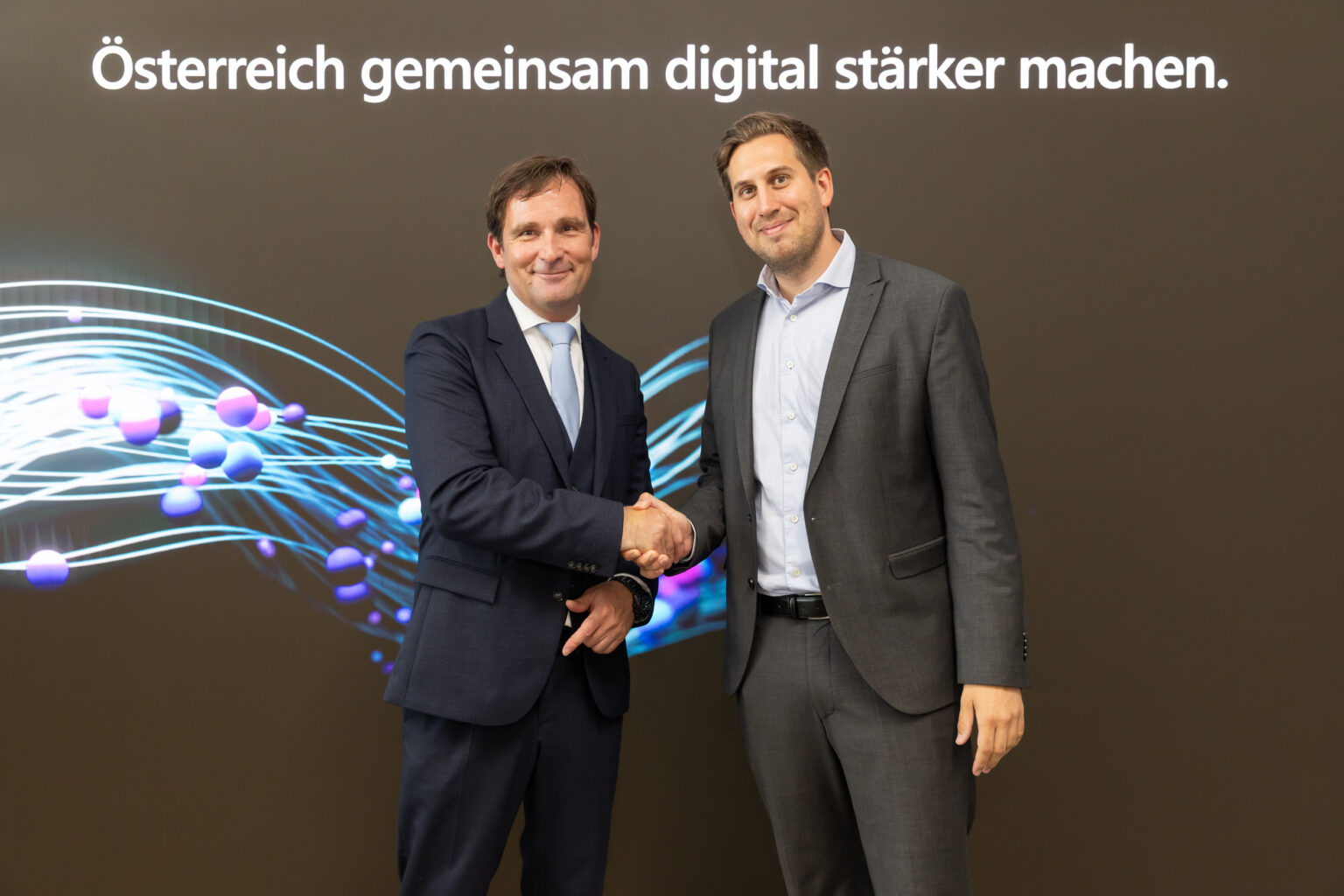 Two men in business suits shake hands and smile at the camera in front of a dark backdrop with the text 