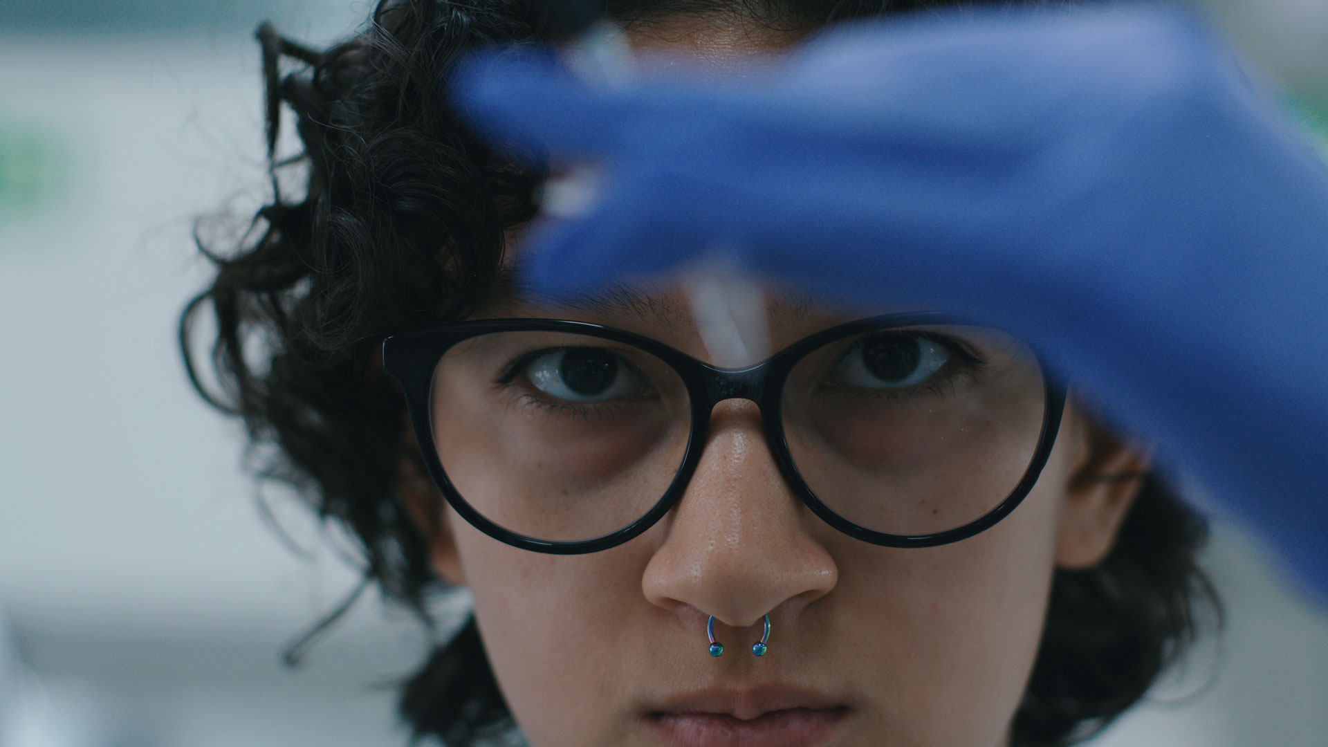 A person with curly hair, glasses, and a blue nose ring focuses intently while holding up a lab instrument, wearing a blue glove in a laboratory setting.