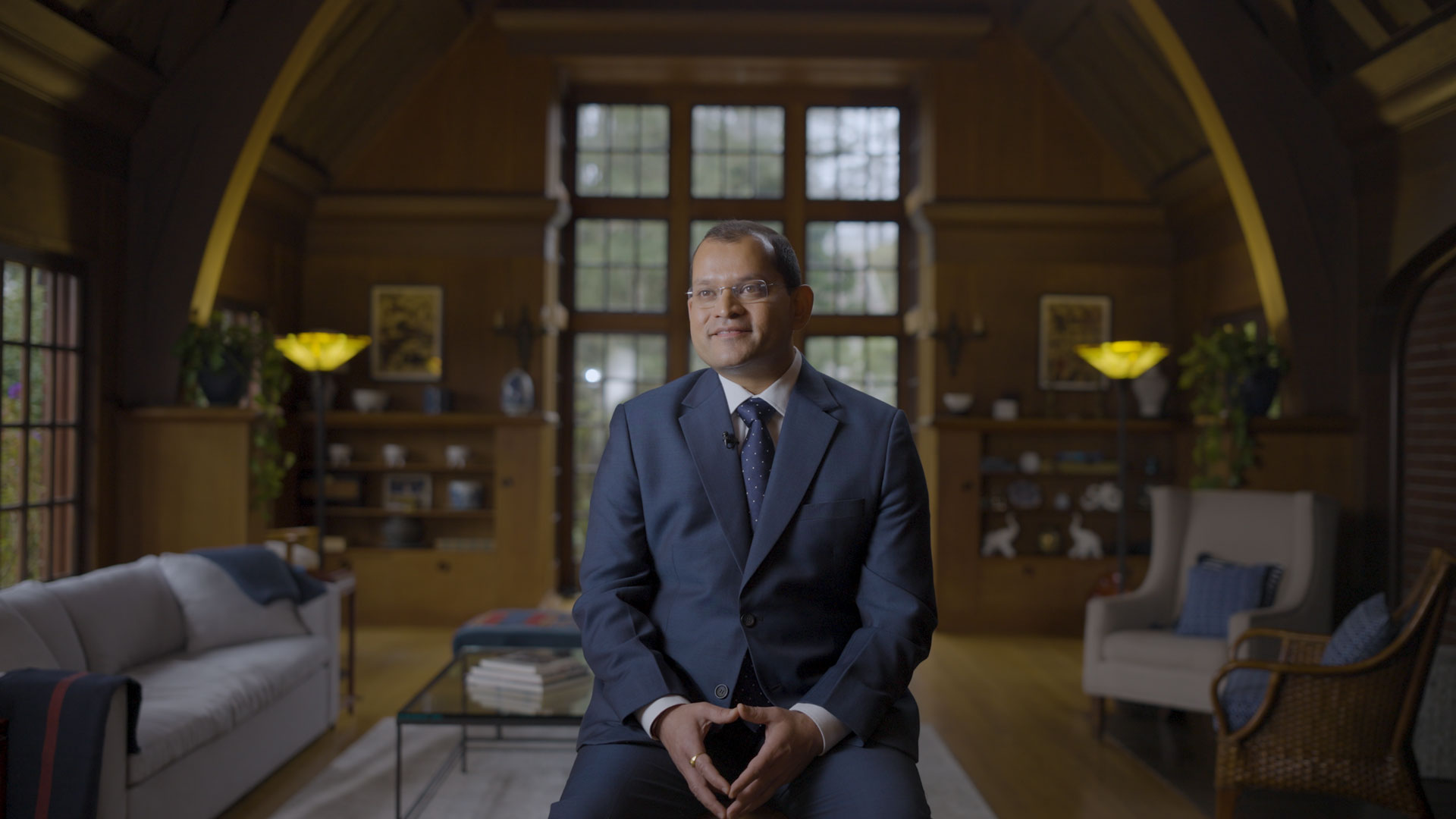A man in a blue suit sits smiling in a well-lit, elegant living room with wooden walls, large windows, bookshelves, and cozy furniture.
