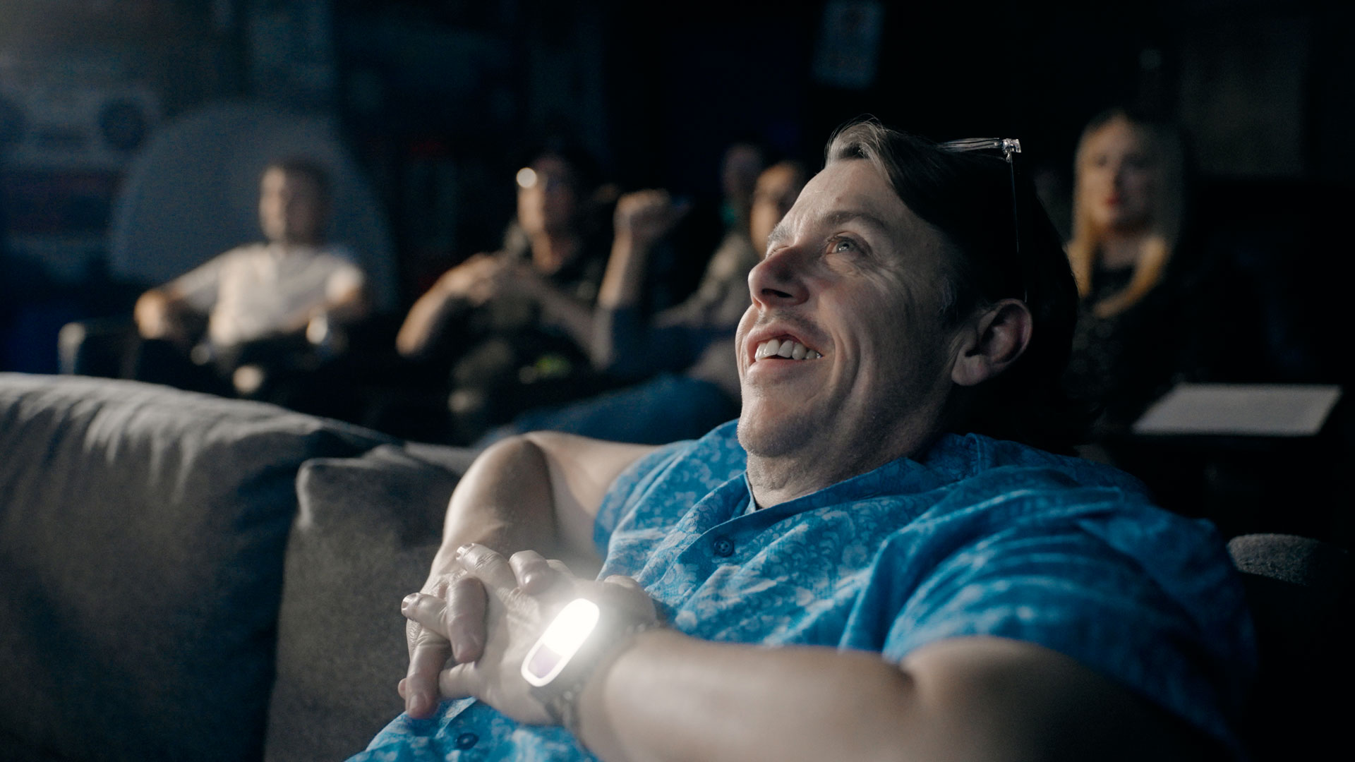 A man in a blue shirt smiles and leans back on a couch, watching something with enjoyment. Several people are seated in the background, also watching and reacting. The setting appears to be a home theater or screening room.