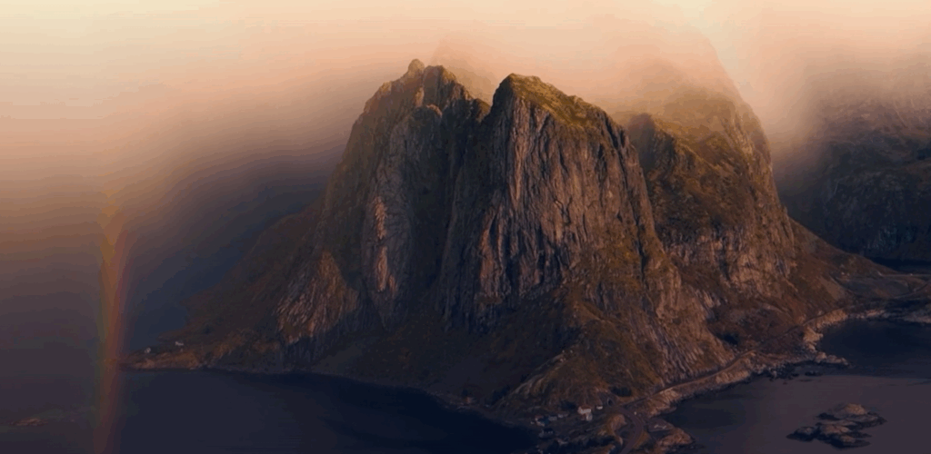 A tall, rocky mountain rises from the sea, partially shrouded in mist and low clouds, with a faint rainbow appearing on the left and soft, warm sunlight illuminating the scene.
