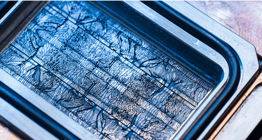 Close-up of a black rectangular printing plate with intricate, abstract line patterns, possibly for printmaking or etching, illuminated by blue-tinted light.