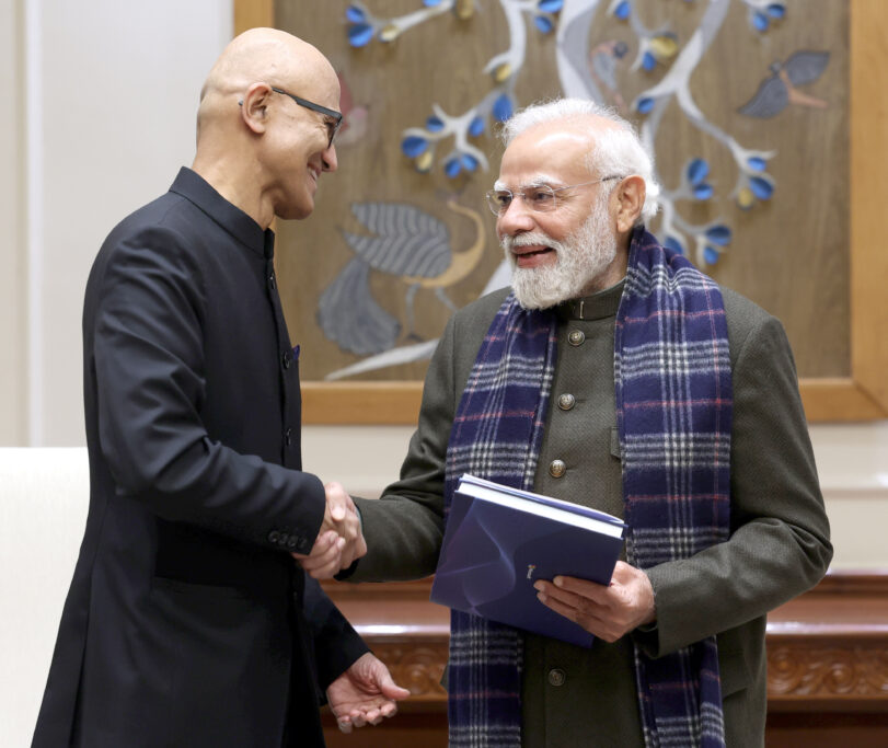 Two men in formal attire shake hands and smile at each other; one holds a folder. They stand in front of a decorative wall with floral artwork.
