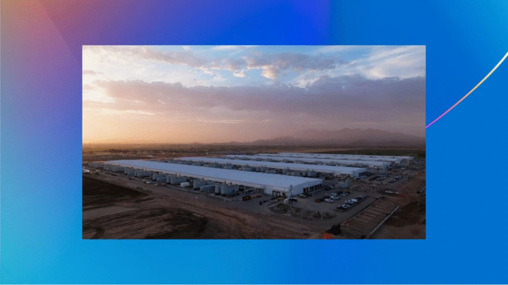 Aerial view of a large industrial facility with multiple long white buildings, set against a sunrise or sunset sky with mountains in the distance, surrounded by parking areas and dirt fields.