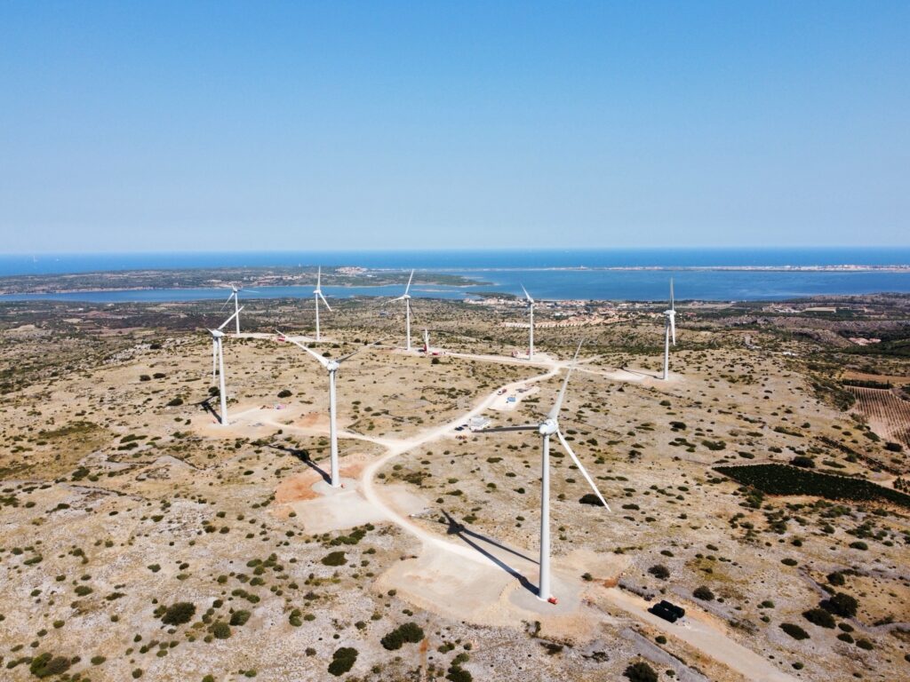Aerial view of a wind farm with multiple wind turbines on a rocky, grassy landscape near the coast, under a clear blue sky with the sea visible in the distance.