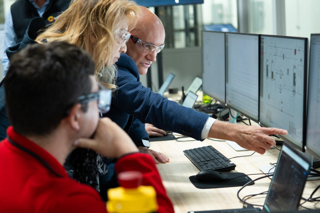 Three people wearing safety glasses look at computer monitors displaying technical diagrams; one person points at a screen, discussing the data, in a modern office or control room setting.