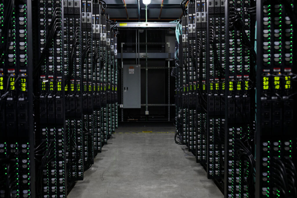 Rows of server racks with glowing lights line both sides of a narrow aisle in a data center, with concrete floors and industrial lighting overhead.