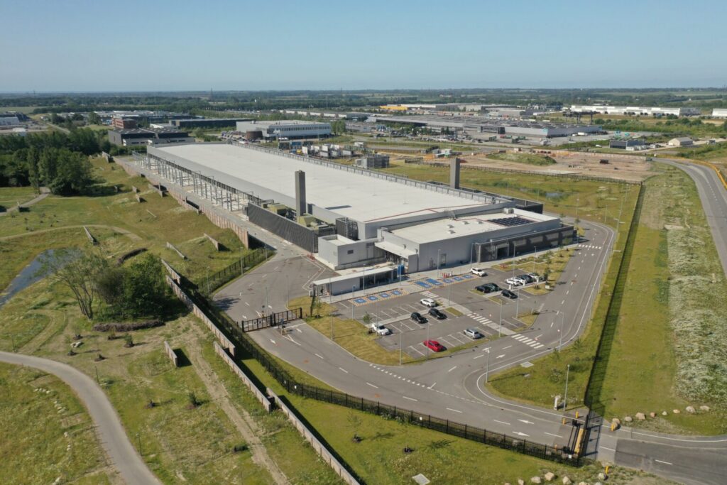 Aerial view of a large industrial warehouse with a white roof, parking lots, and surrounding roads on a clear day. Some vehicles are parked, and greenery surrounds the facility. Other buildings are visible in the distance.