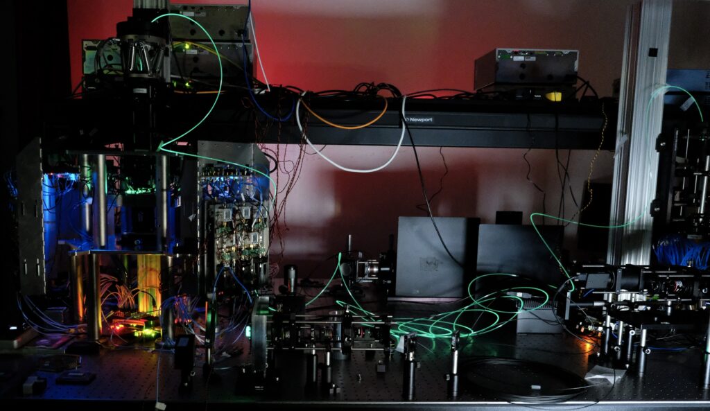A laboratory setup with various electronic devices, wires, and glowing green laser beams running across optical equipment on a metal table, all illuminated in dim lighting.