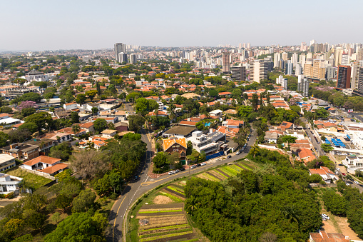 Aerial view of a city with a mix of tall modern buildings and smaller houses, surrounded by green trees and a park with gardens, under a clear sky.