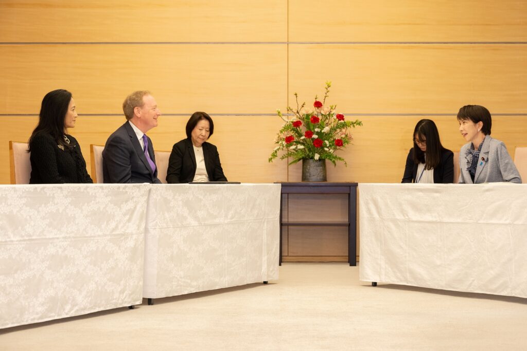 Five people in formal attire sit across from each other at two tables with white tablecloths, engaged in conversation. A floral arrangement is displayed on a table against a wooden wall in the background.
