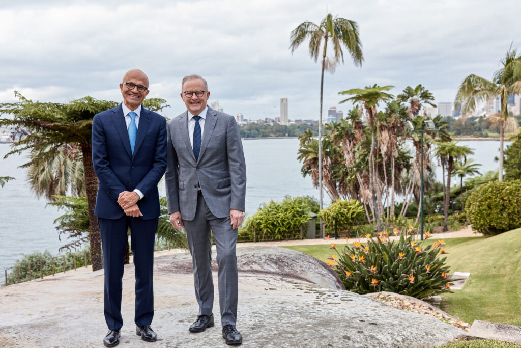 Two men in suits stand smiling outdoors on a stone pathway, with lush greenery, palm trees, and a city skyline in the background across the water under a cloudy sky.