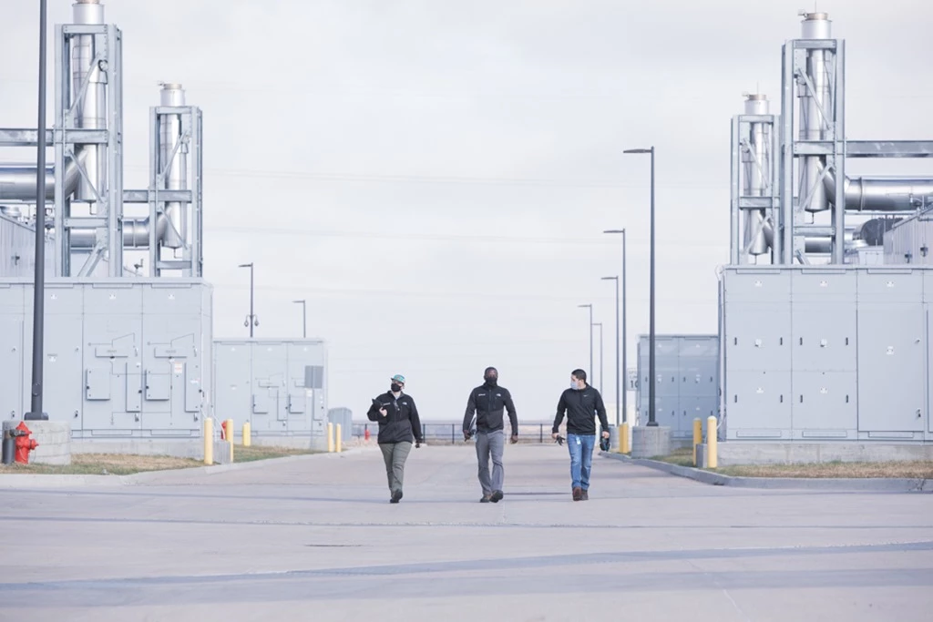 Three people walk down a wide paved roadway between two large industrial buildings with metal structures and equipment on a cloudy day.