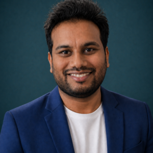 A man with short dark hair and a beard smiles at the camera. He is wearing a white shirt and a blue blazer, standing in front of a dark teal background.
