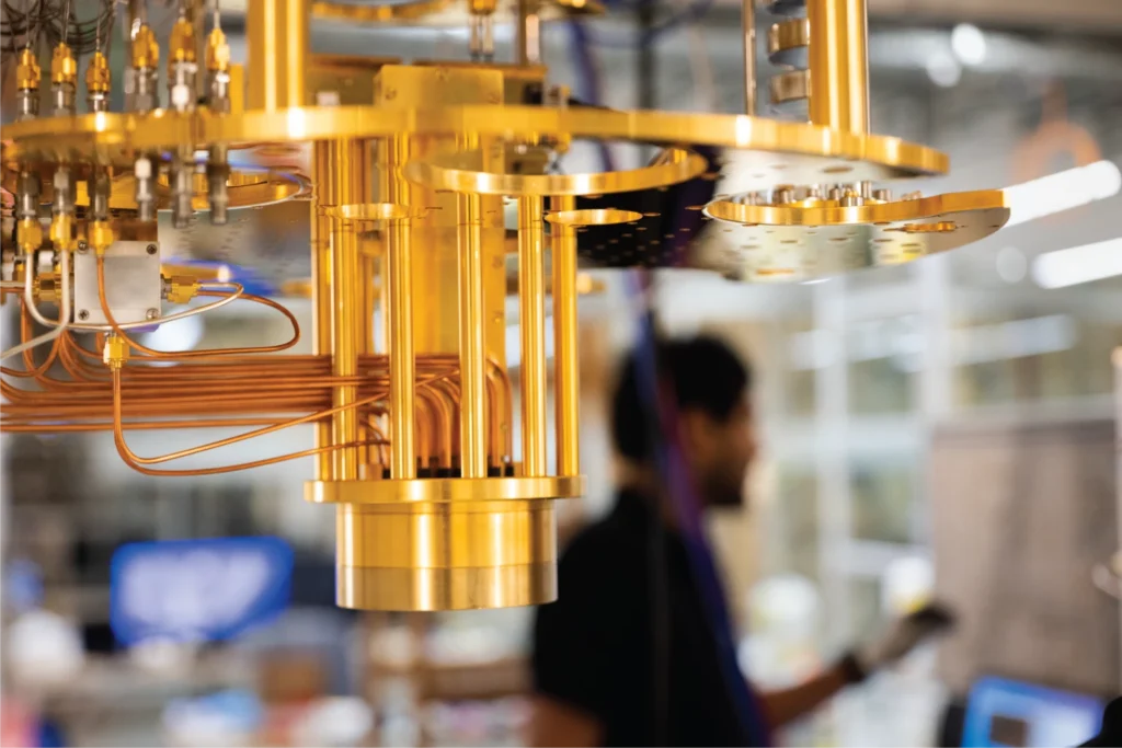 A close-up of a gold-colored quantum computer component, showing wires and circuitry, with a blurred person and laboratory background.