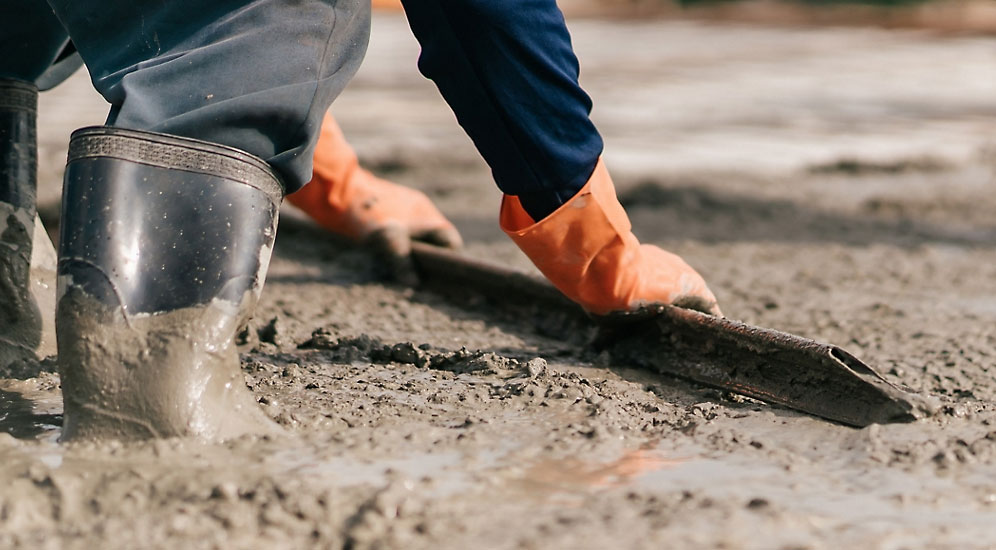 Close-up of a construction worker wearing rubber boots and orange gloves, smoothing wet concrete with a leveling tool on a construction site.