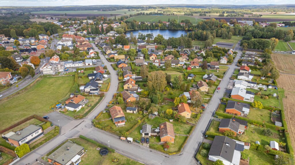 Aerial view of a small town with houses, roads, greenery, and a pond in the background, surrounded by fields and trees on a clear day.
