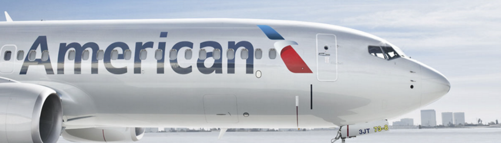 A close-up side view of an American Airlines airplane on the ground, with the logo and part of the fuselage visible. City buildings and a cloudy sky appear in the background.