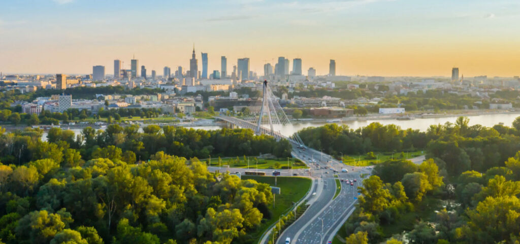 Aerial view of a city skyline at sunset with a modern bridge spanning a river, surrounded by lush green trees, roadways, and tall buildings in the background.