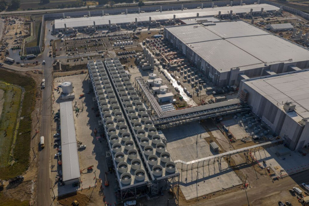 Aerial view of a large industrial facility under construction, featuring rows of large cooling fans, multiple warehouse-like buildings, parked vehicles, and surrounding roads.