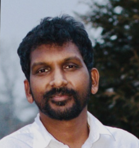 A man with short curly hair and a beard, wearing a white shirt, stands outdoors with trees and a cloudy sky in the background.