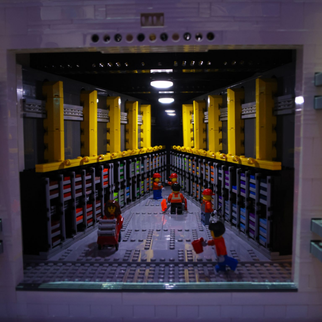 A LEGO model depicts a data center with yellow racks, black and colorful server units, and four LEGO minifigures in orange vests working inside the aisle under bright ceiling lights.