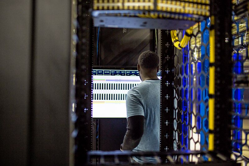 A person stands in a server room, facing multiple computer monitors displaying data, surrounded by server racks and network cables.