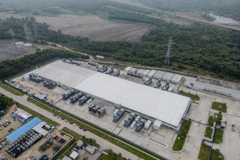 Aerial view of a large industrial facility with a white roof, surrounded by trees and fields, with several machines and structures around the building and high-voltage power lines nearby.