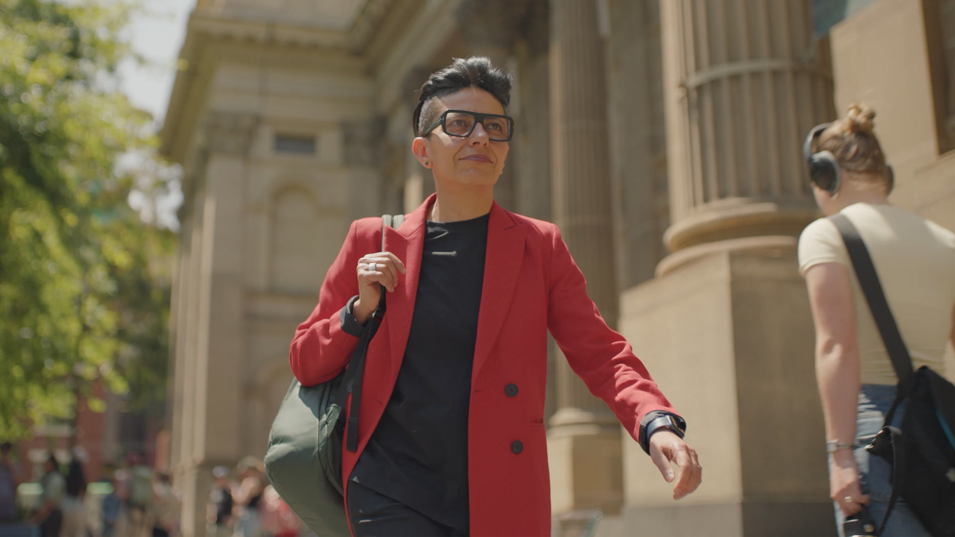 A person wearing a red blazer, black shirt, and glasses walks confidently outside a large stone building, carrying a green bag. Another person with headphones and a backpack is seen in the background.