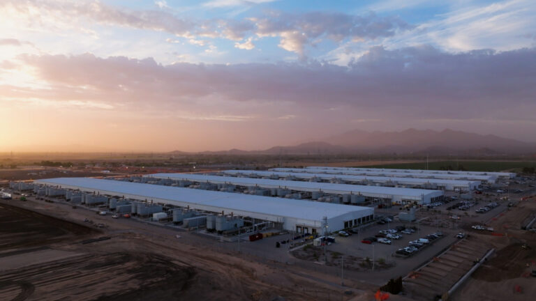 A large industrial facility with multiple long, white-roofed buildings and a parking lot sits in a desert landscape at sunset, with mountains and a partly cloudy sky in the background.