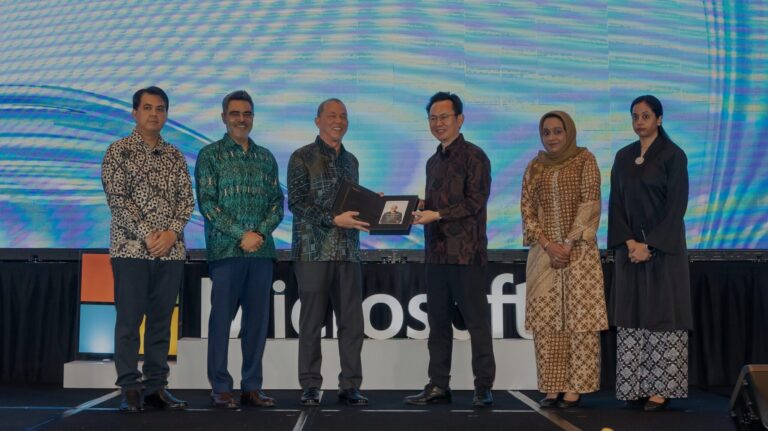 Six people stand on a stage in front of a screen displaying the Microsoft logo. Two men in the center are shaking hands while holding a plaque. The group is dressed in formal and traditional attire.