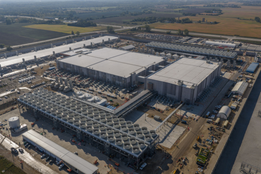Aerial view of a large data center complex surrounded by fields, featuring multiple rectangular buildings, cooling towers, construction equipment, and vehicles on site.