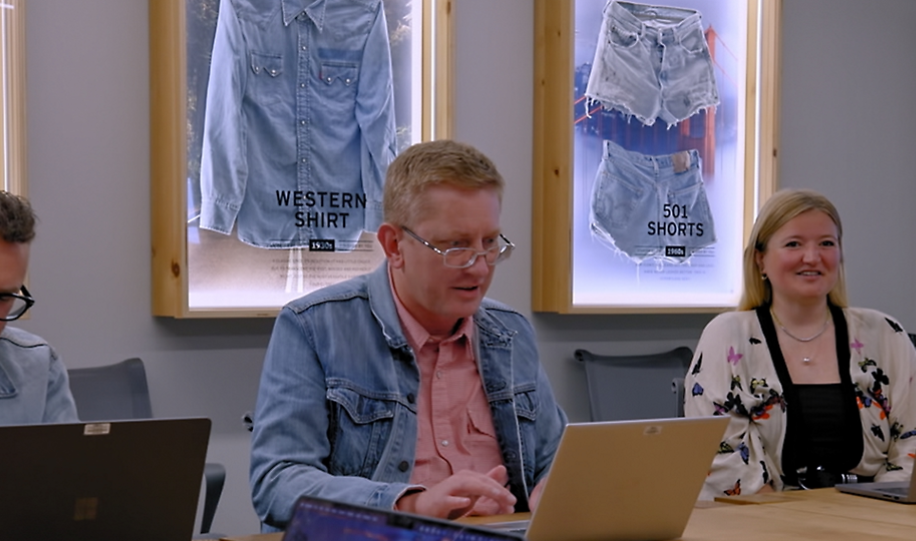 Three people sit at a table with laptops in a meeting room. Behind them are framed displays of denim shirts and shorts on the wall. One man in glasses types on a laptop, while a woman smiles beside him.