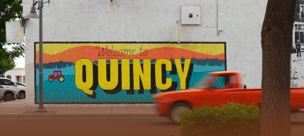 A mural on a white wall reads “Welcome to QUINCY” with a small red tractor. An orange truck drives by in the foreground on a street lined with trees and parked cars.