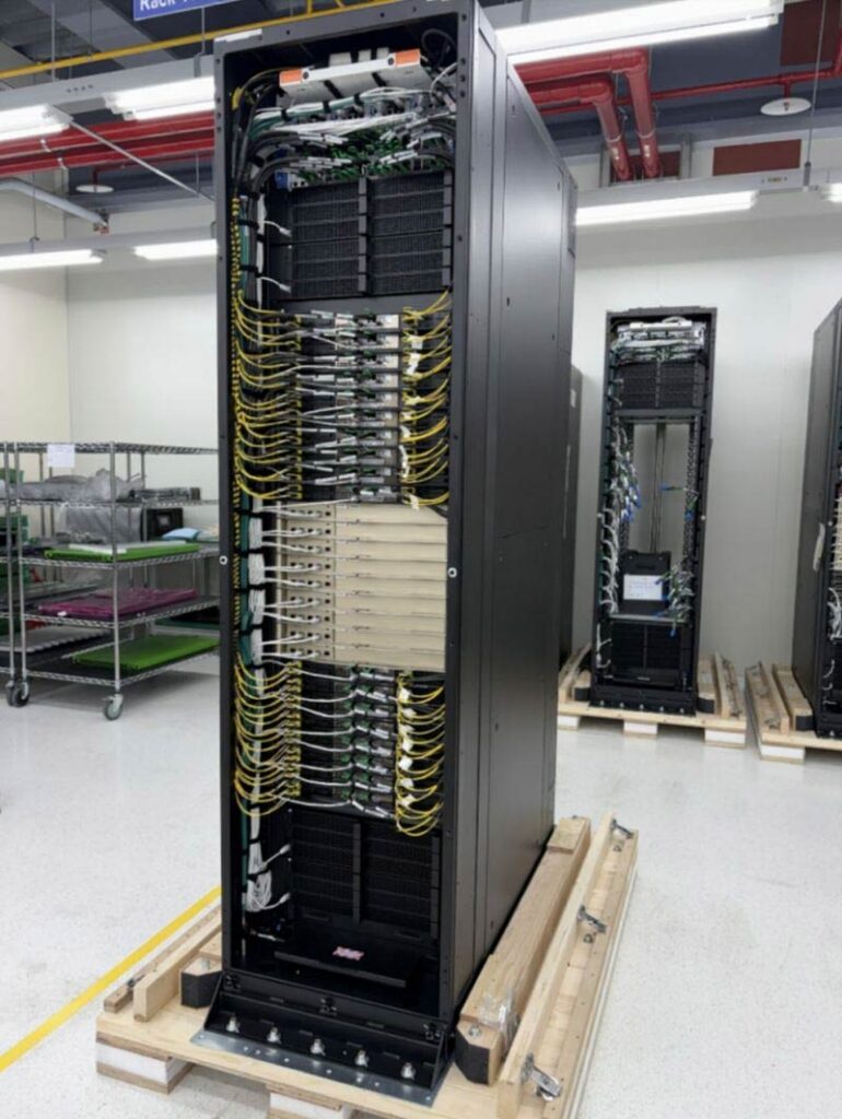 A black server rack filled with networking equipment and organized yellow and white cables stands in a clean, industrial room with other racks and metal shelves visible in the background.