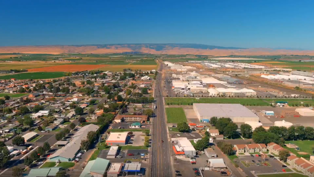 Aerial view of a small town with a main road running through it, fields and industrial buildings on either side, and distant hills under a clear blue sky.