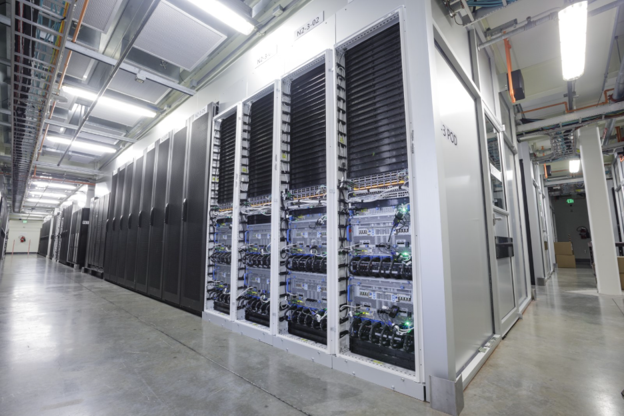 Rows of server racks with visible cables and glowing indicator lights are arranged in a brightly lit, modern data center with a clean, polished concrete floor.