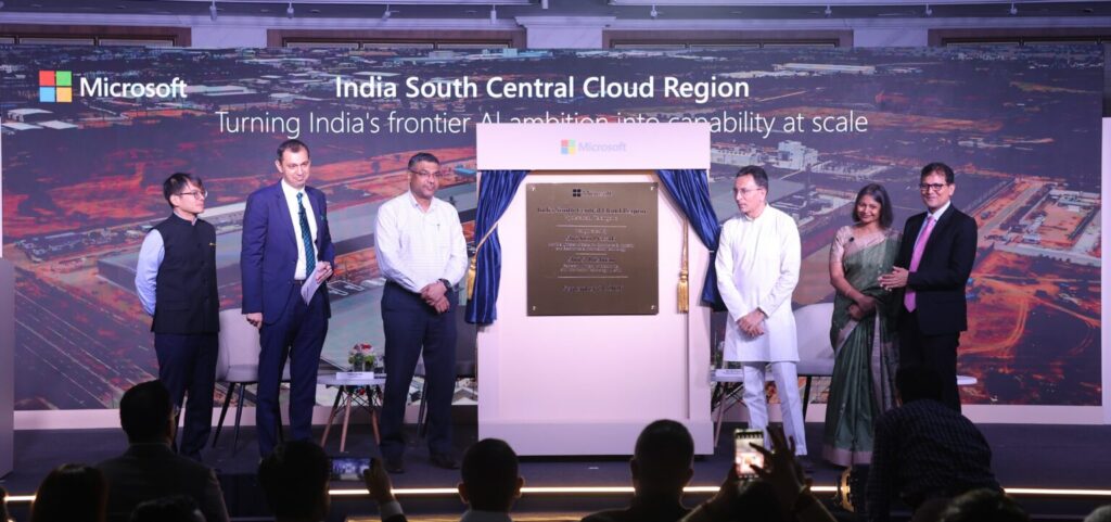 Six people stand on stage beside a plaque at a Microsoft event for the India South Central Cloud Region, with a backdrop showing event details and a cityscape image.