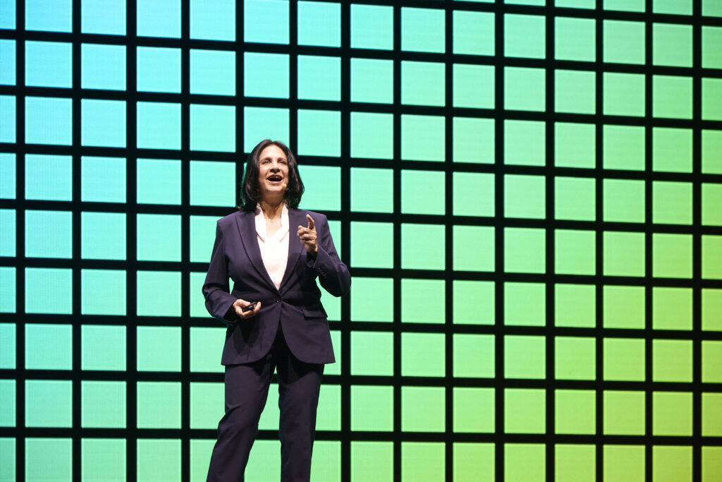 A woman in a dark suit stands and gestures while speaking on stage in front of a large green-and-blue grid background.