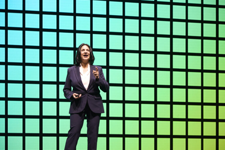 A woman in a dark suit stands and gestures while speaking on stage in front of a large green-and-blue grid background.