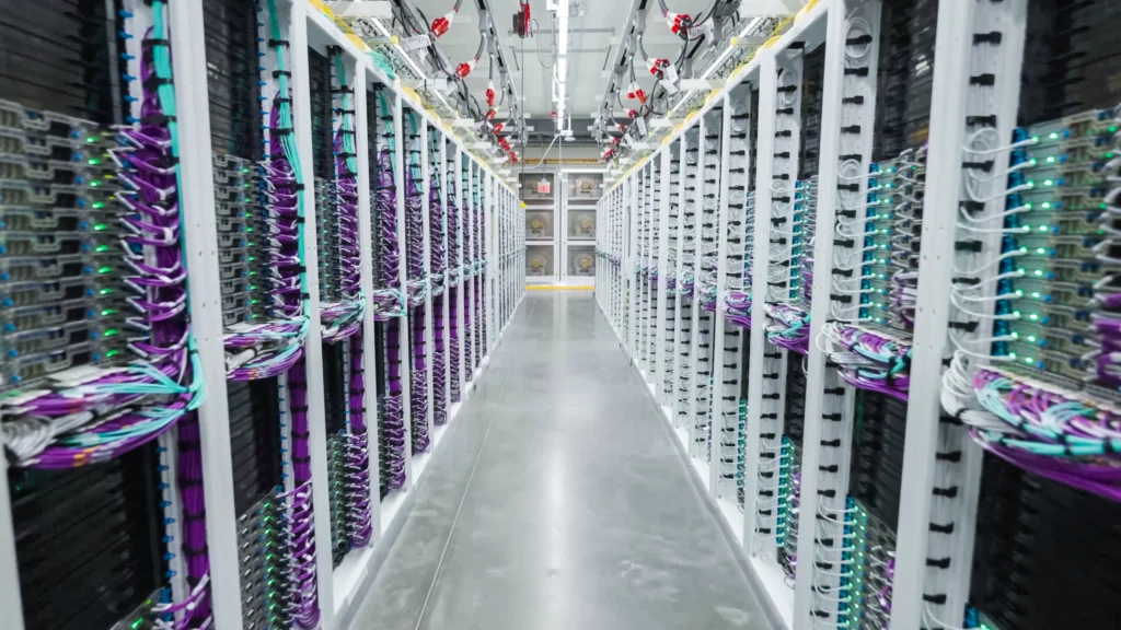 A narrow aisle between two rows of server racks with glowing indicator lights and bundles of purple and teal network cables.